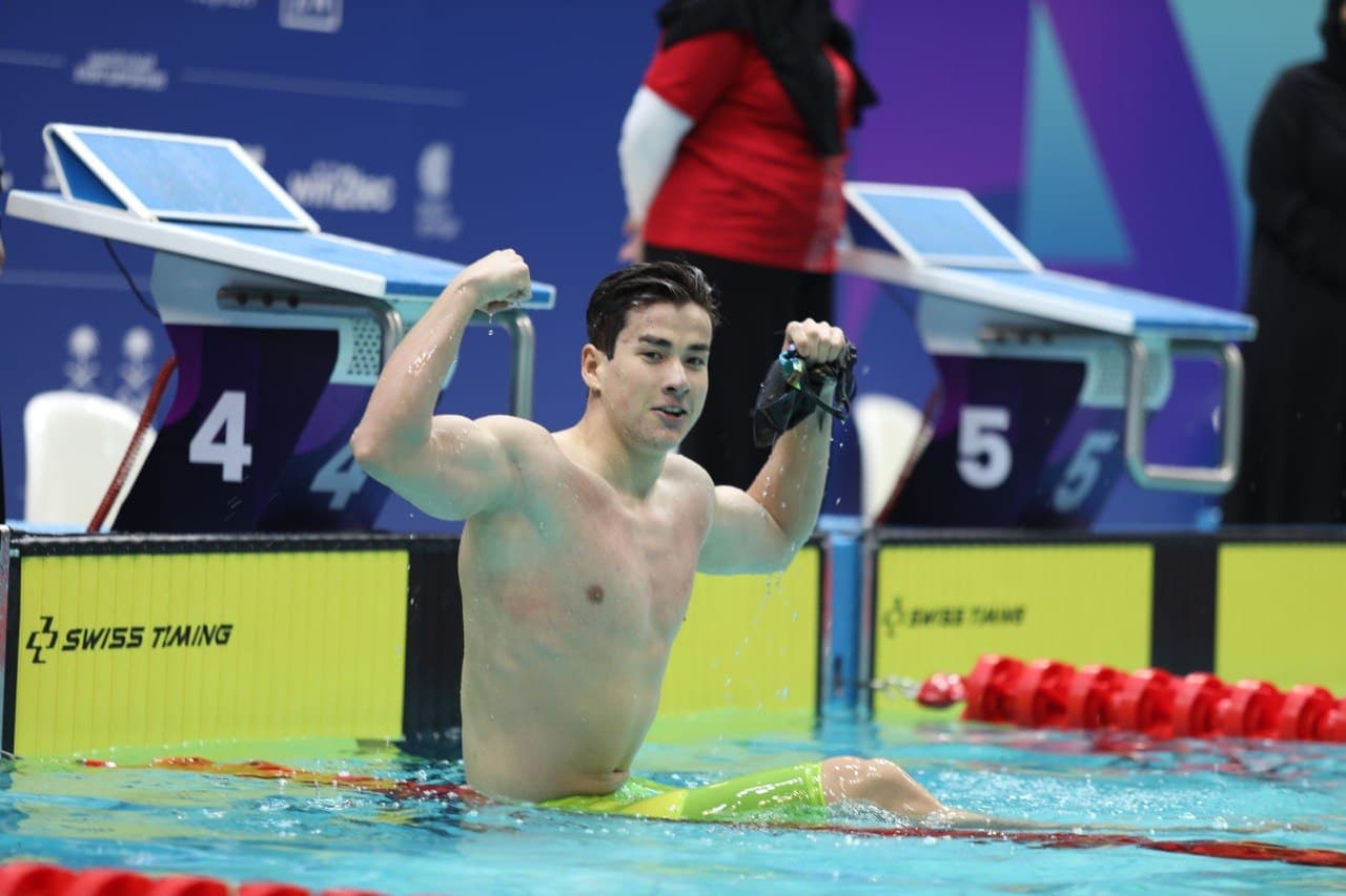 Male swimmer celebrates in a pool, flexing his bicep and holding goggles near starting blocks.