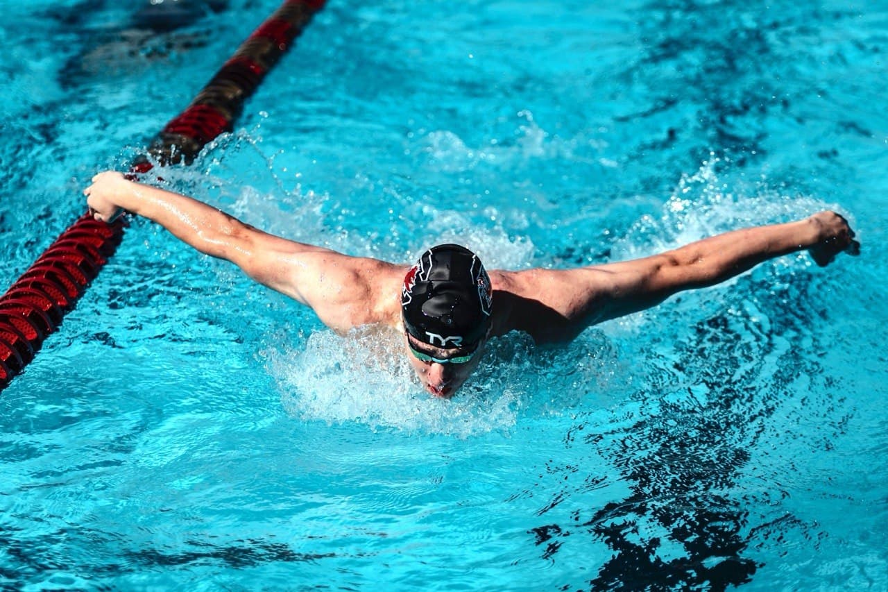 Swimmer in a black cap performs the butterfly stroke, splashing through bright blue pool water.